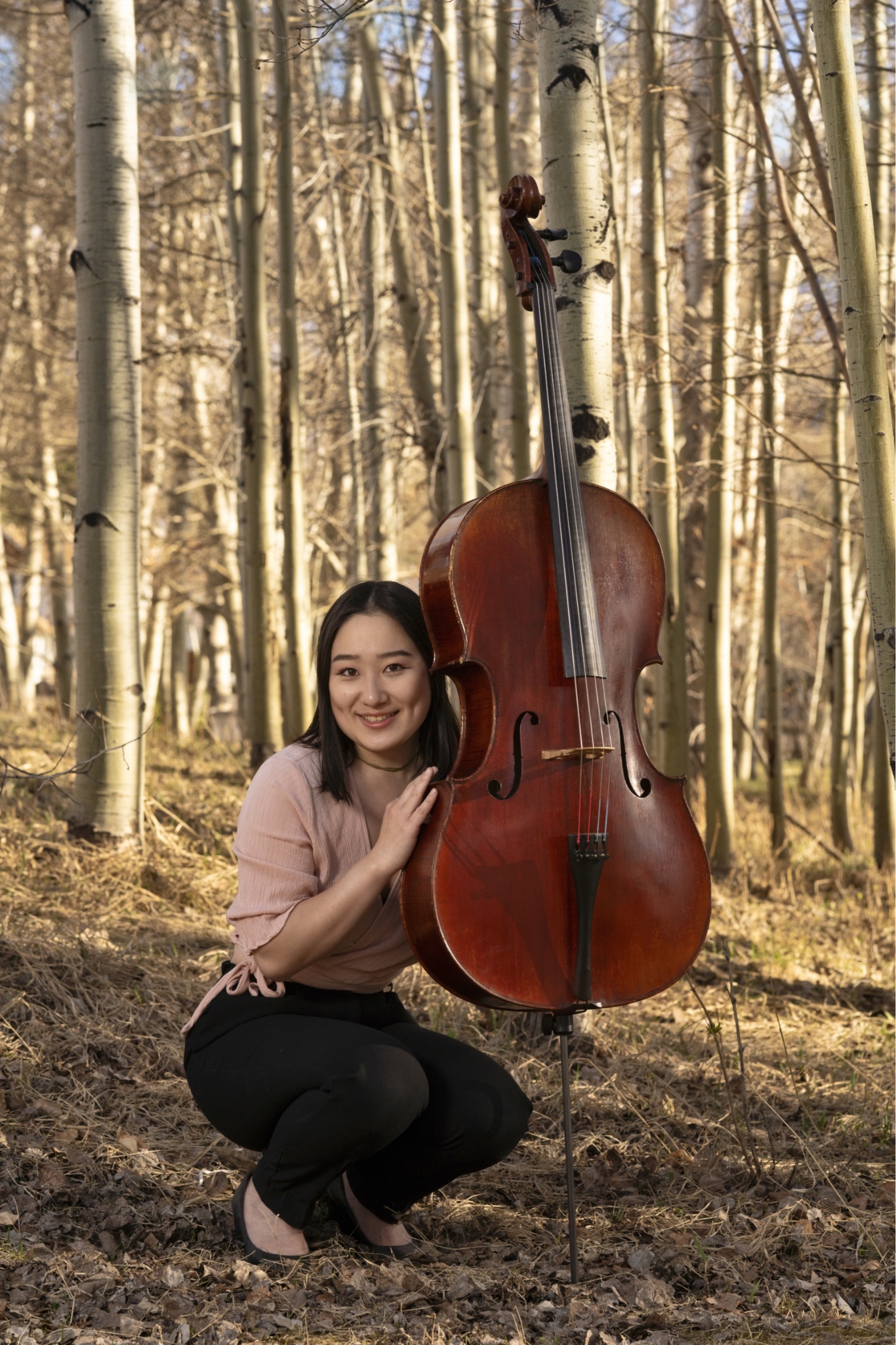 Erica Ogihara crouching with her cello in the woods